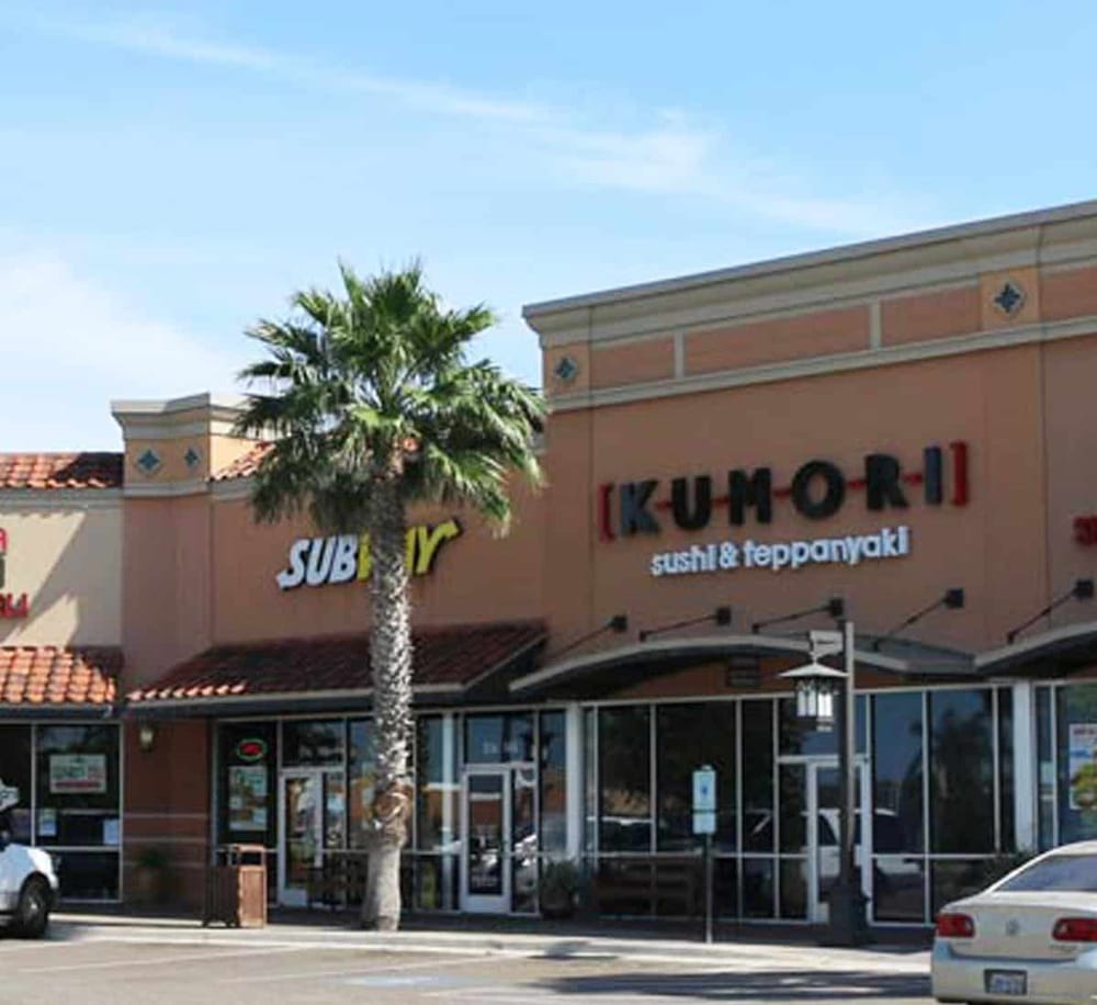 Vibrant shopping center with Kumori sushi bakery and Subway restaurant, palm tree in front, clear blue sky.
