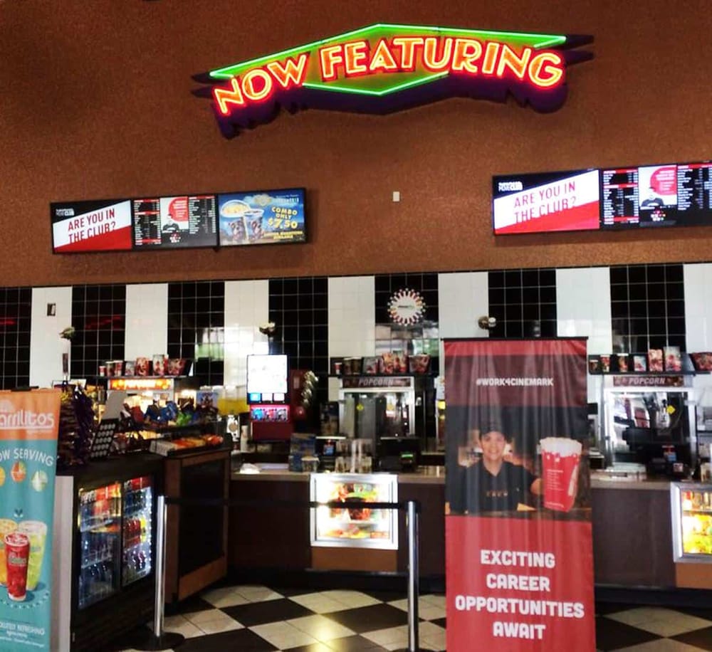 Neon "Now Featuring" sign at a cinema concession stand with electronic menu boards.