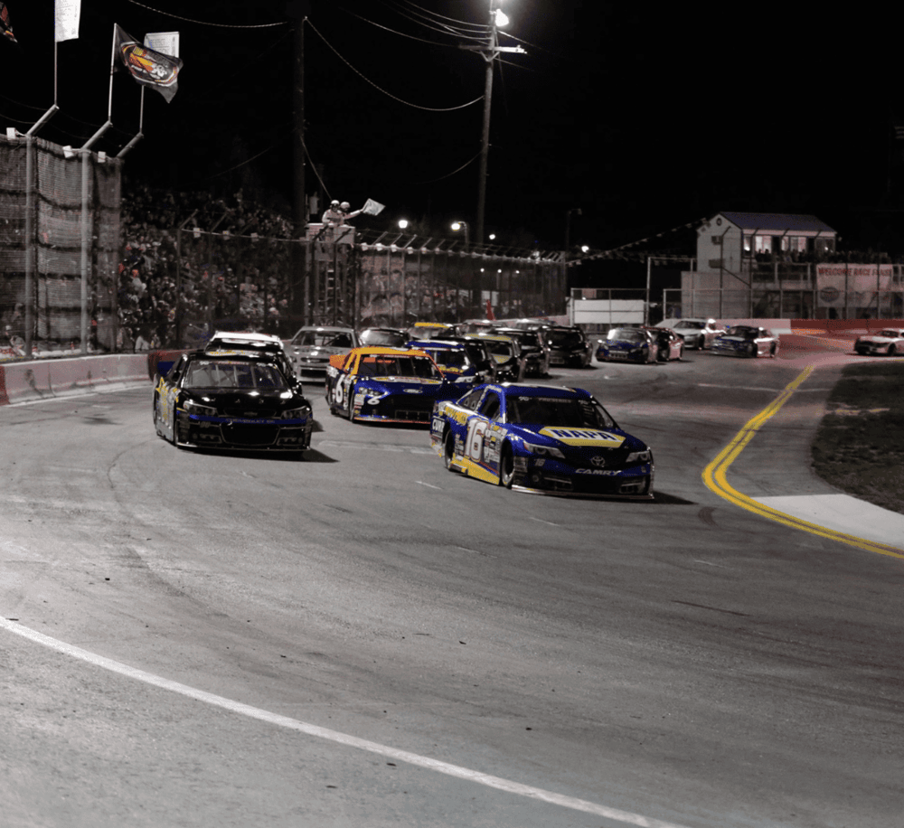 Race car night race at QuestForDirections Speedway with NASCAR vehicles, bright lights, and cheering fans.