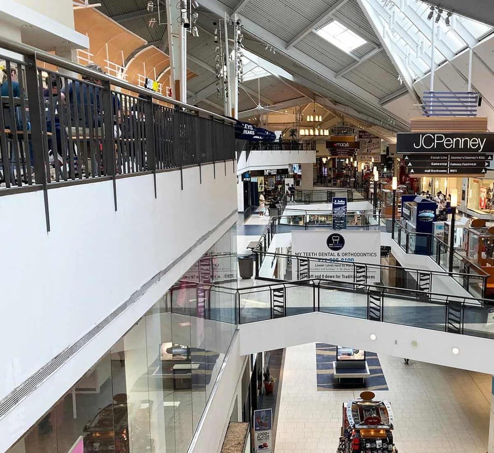 Bright indoor shopping mall with multiple levels, stores, and escalators.