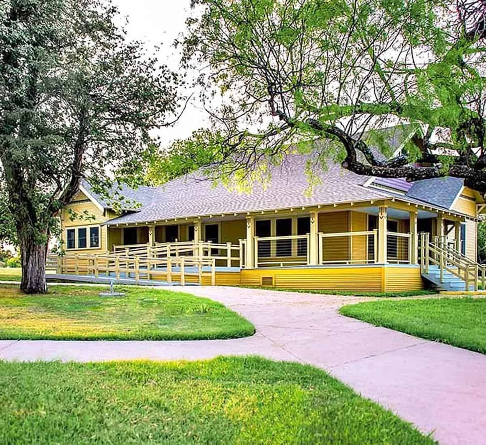 Barefoot walking path leading to a charming yellow house with a spacious porch, surrounded by lush greenery and mature trees, in a peaceful suburban neighborhood.