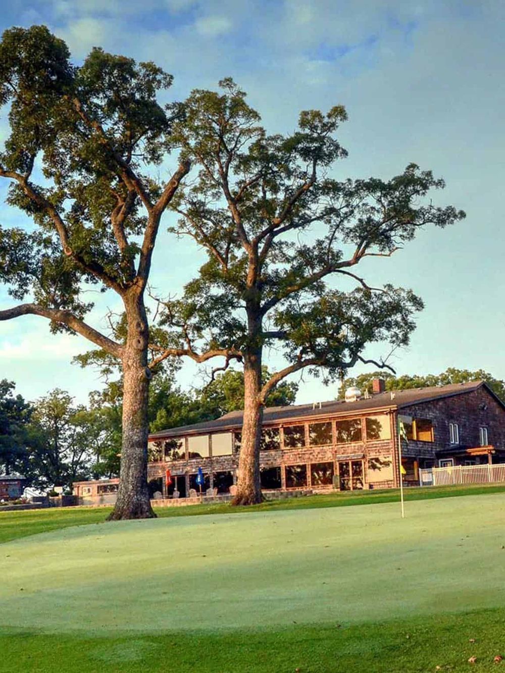 Lush green golf course with large trees and a modern clubhouse under a blue sky.