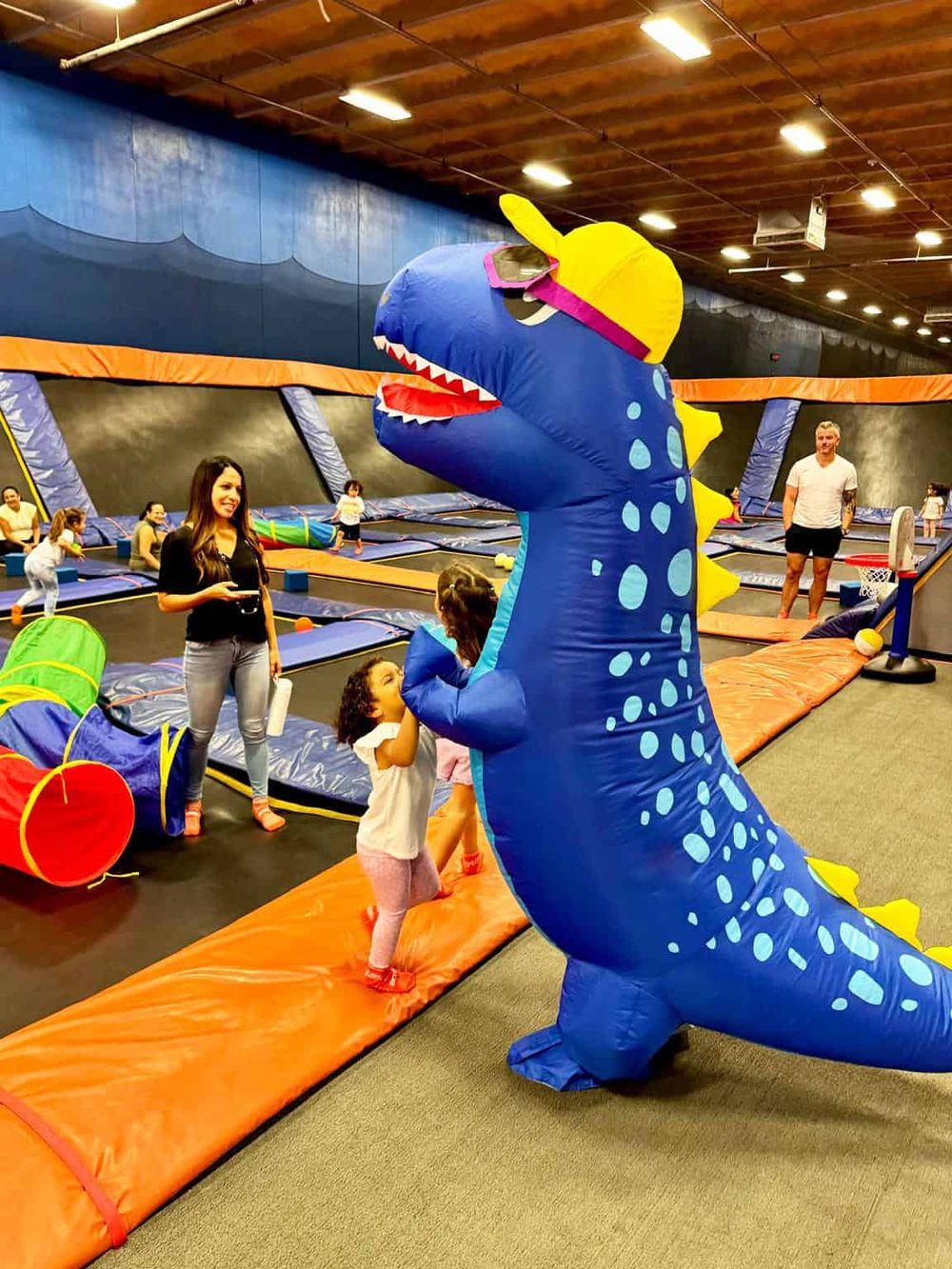 Colorful dinosaur mascot entertaining children at indoor trampoline park.