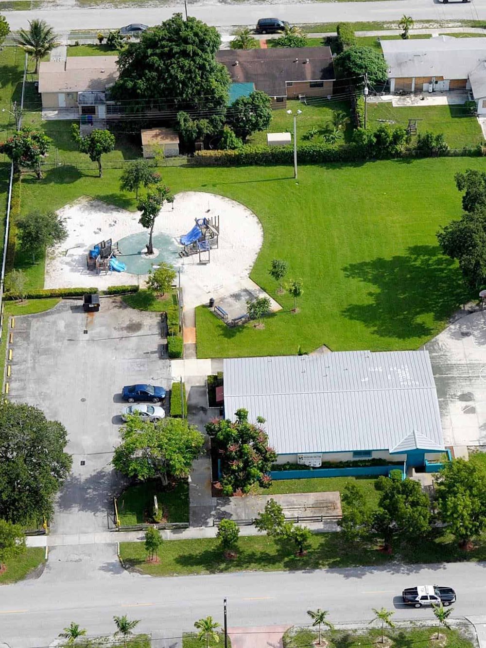 Aerial view of a park with playground, green lawn, trees, parking lot, and surrounding houses, showcasing community outdoor space.
