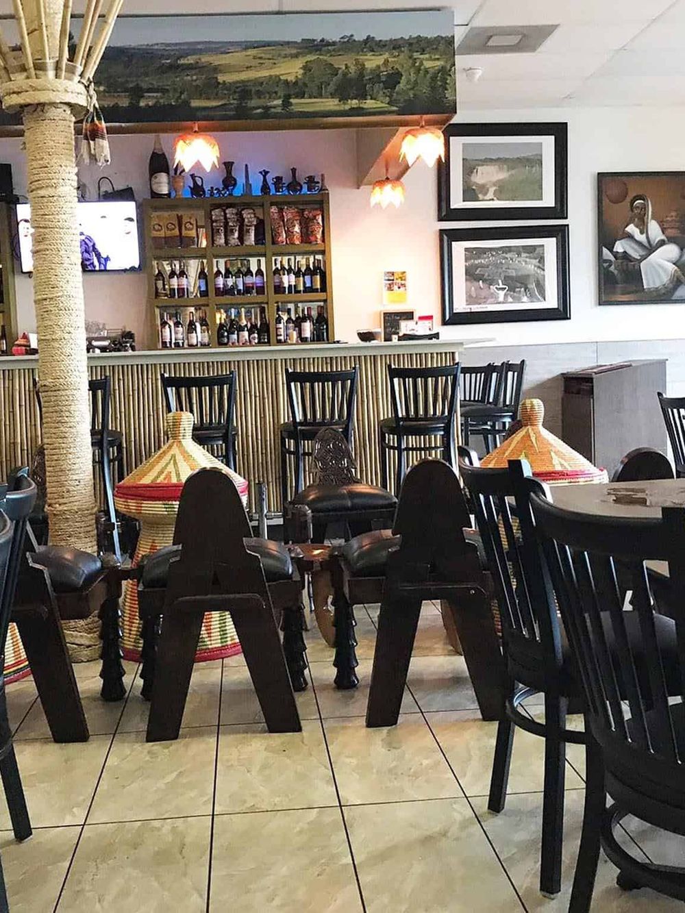 Cozy restaurant interior with bar, black chairs, and colorful woven seating, featuring tropical decor and framed artwork.