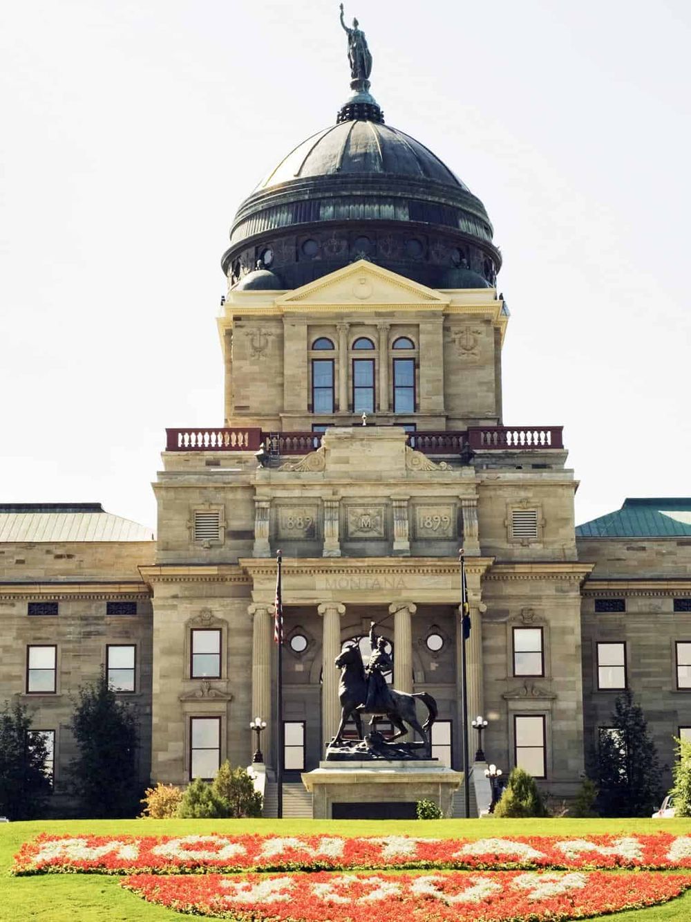 Historic state capitol building in Montana with statue and vibrant flower bed.