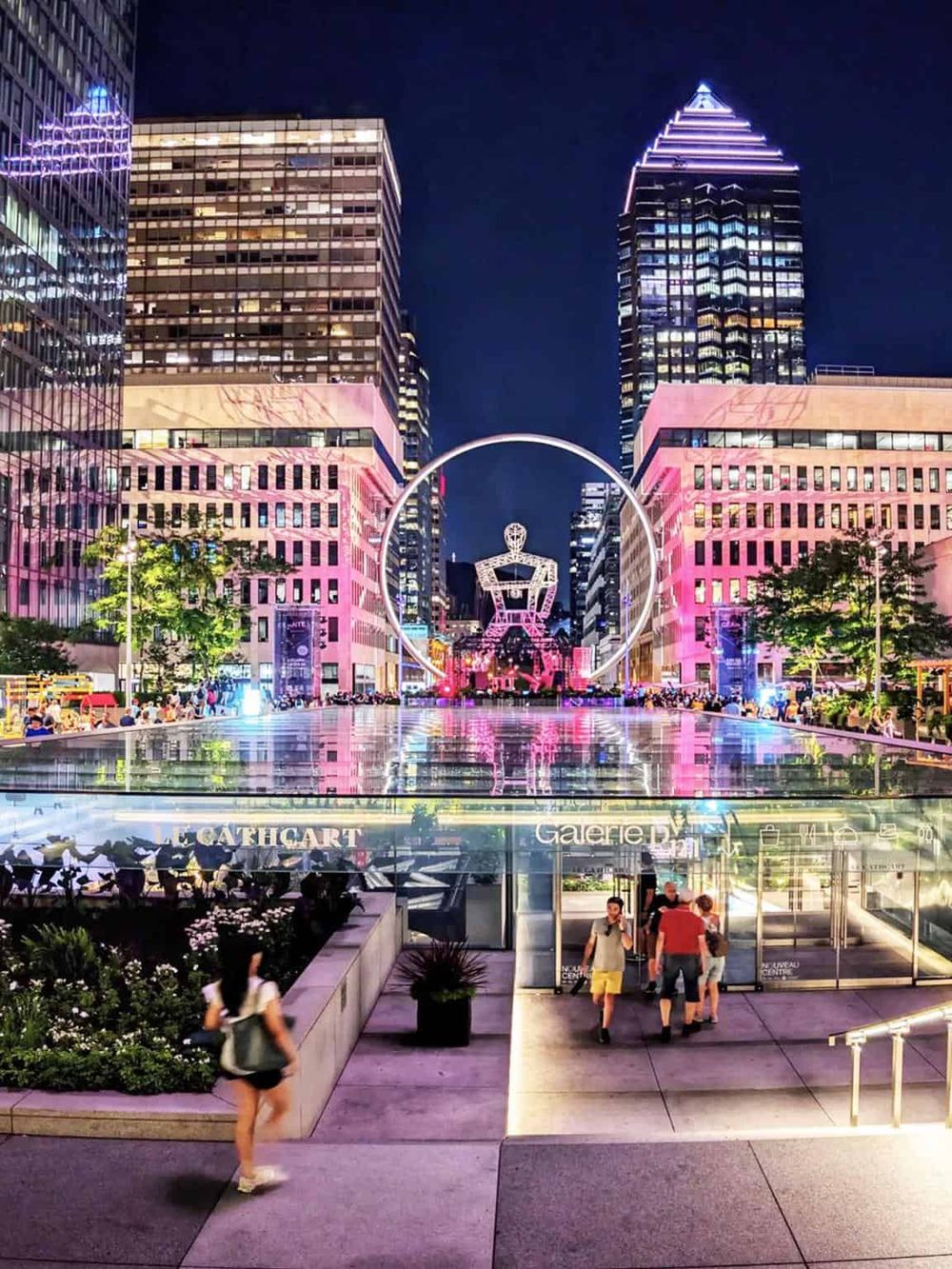 Vibrant night view of downtown with illuminated skyscrapers, modern art installation, and bustling crowd in a cityscape.