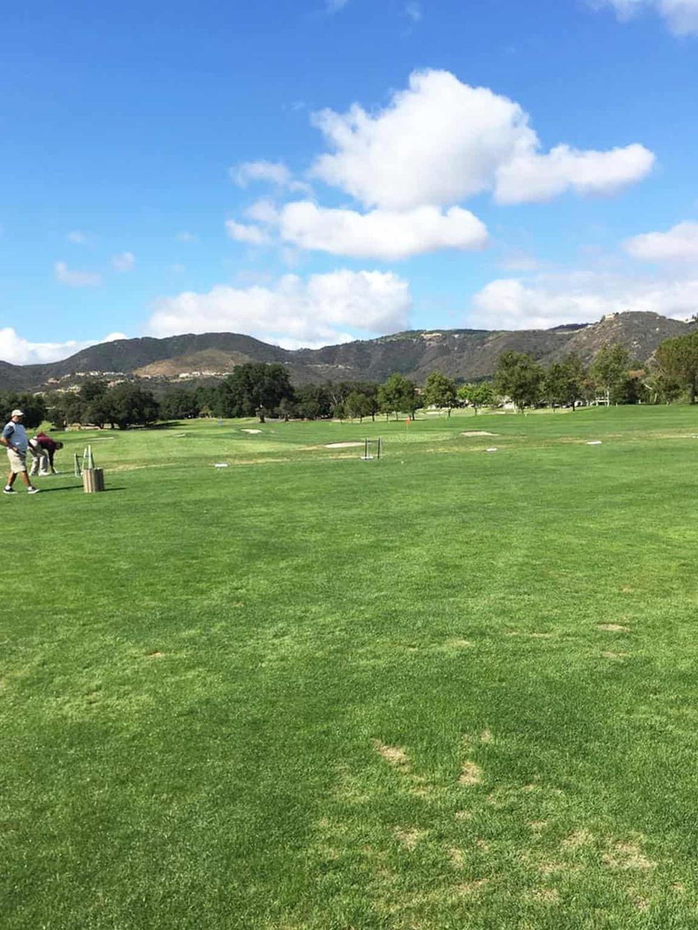 Open outdoor golf course with lush green grass, mountains, and partly cloudy sky in the background.