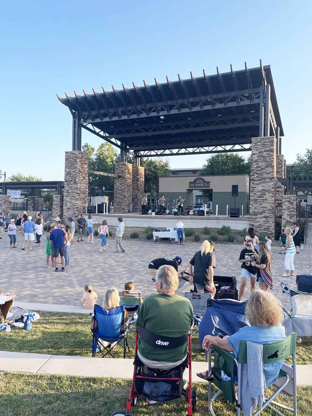 Outdoor live music event with people enjoying a concert on a stage with a canopy in a park setting.