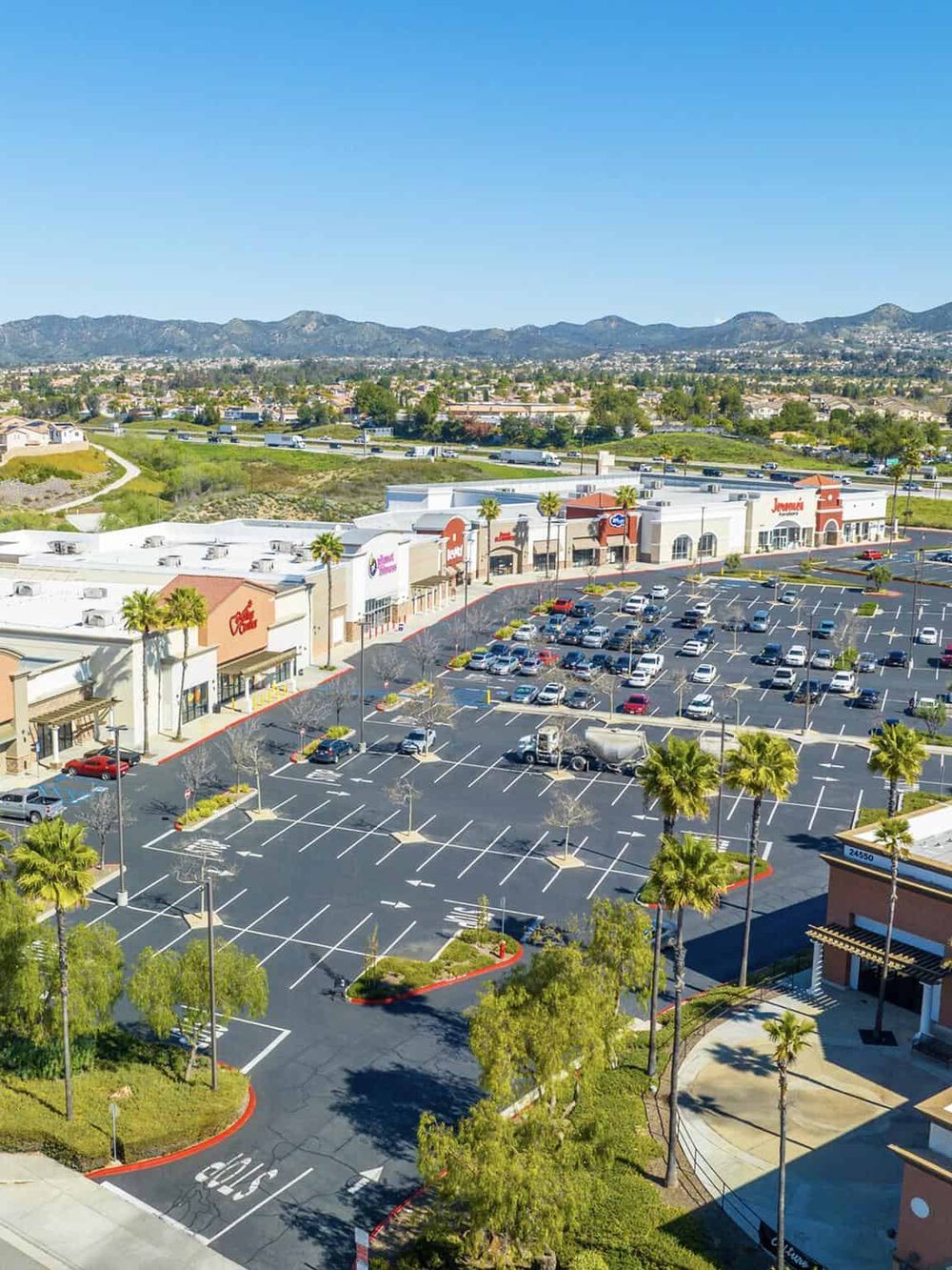 Spacious shopping mall parking lot with retail stores and scenic mountain backdrop.