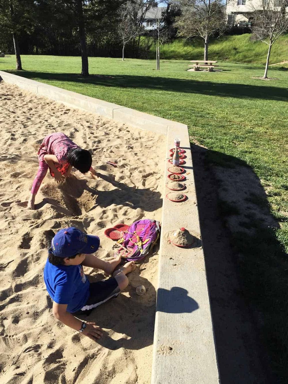 Children playing in a sandbox at a park with greenery and picnic tables in the background.