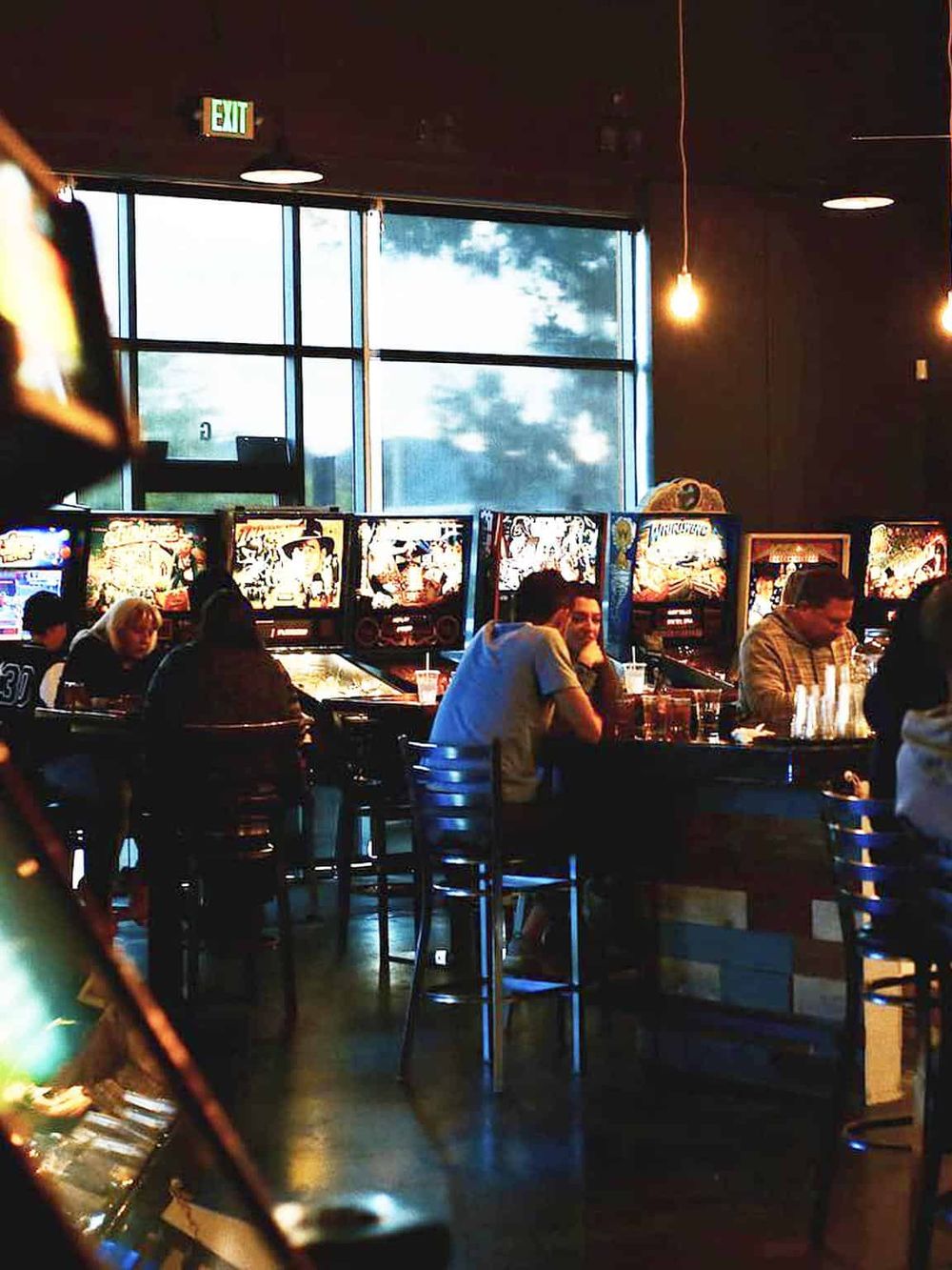 Bright arcade gaming area with pinball machines, patrons enjoying games, modern decor, and large windows letting in natural light.