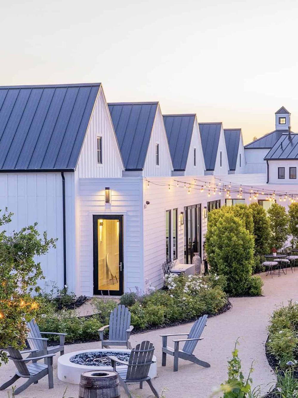 Charming white barn-style venue with outdoor seating and string lights at dusk. Perfect for events and gatherings.