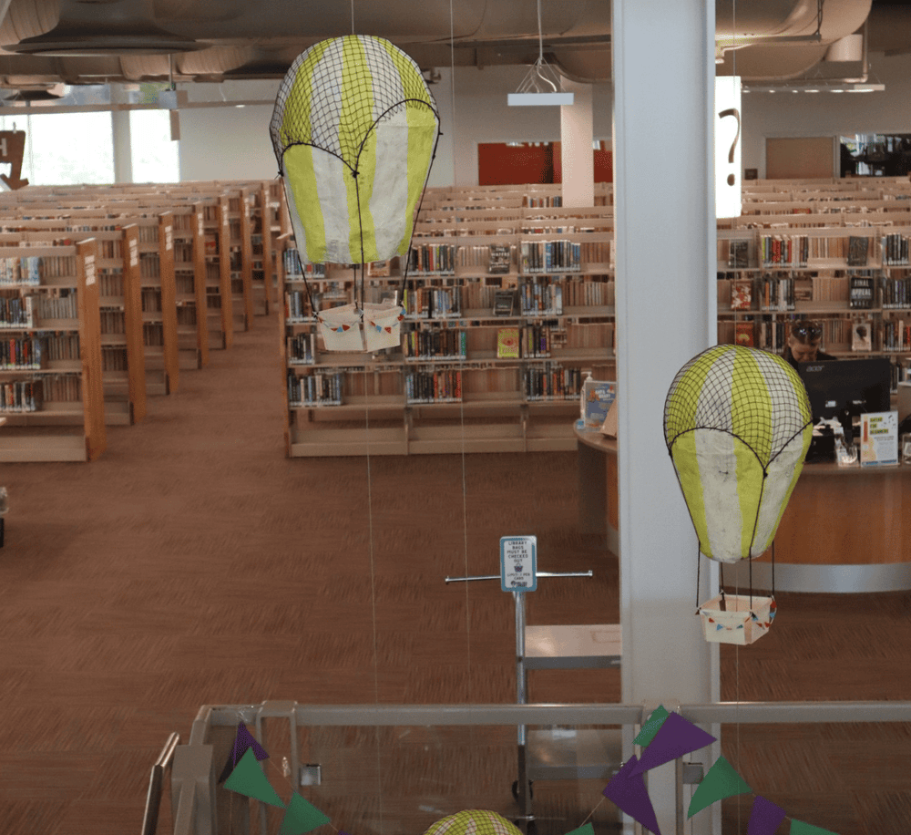 Colorful hot air balloon paper lanterns inside a modern library with wooden bookshelves.