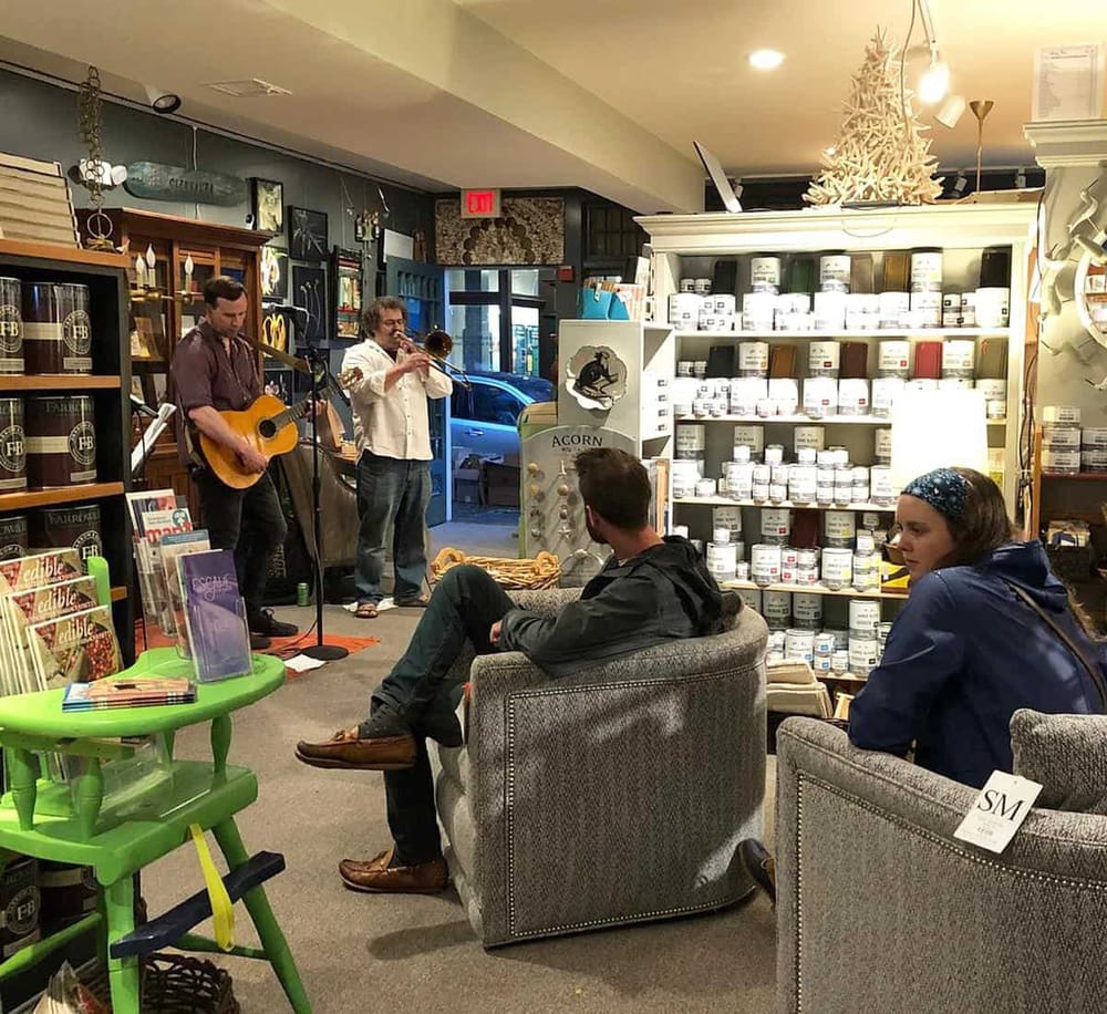 Cozy coffee shop with live music performance, featuring two musicians playing guitar and trumpet, surrounded by customers and artistic decor.