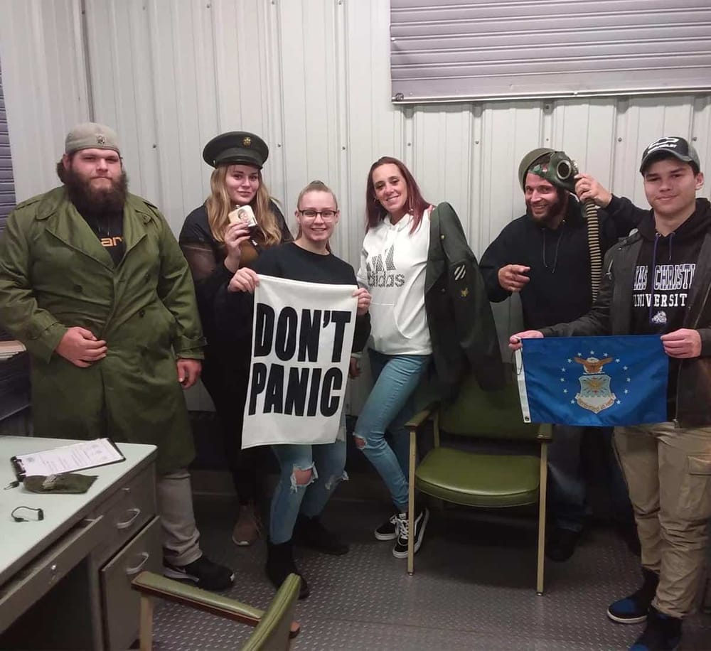 Older man with a beard in military-style green coat and a beanie, standing next to young woman holding a "DON'T PANIC" sign, group photo.