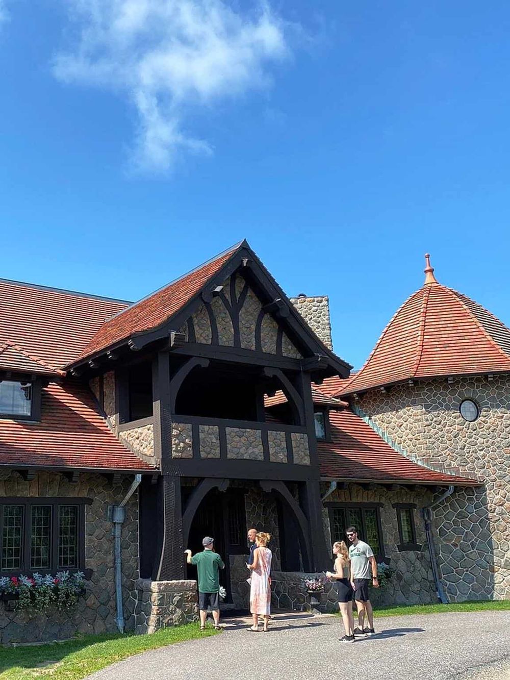 Historic house with stone walls and red-tile roof, featuring a black timber-framed balcony, under a bright blue sky.