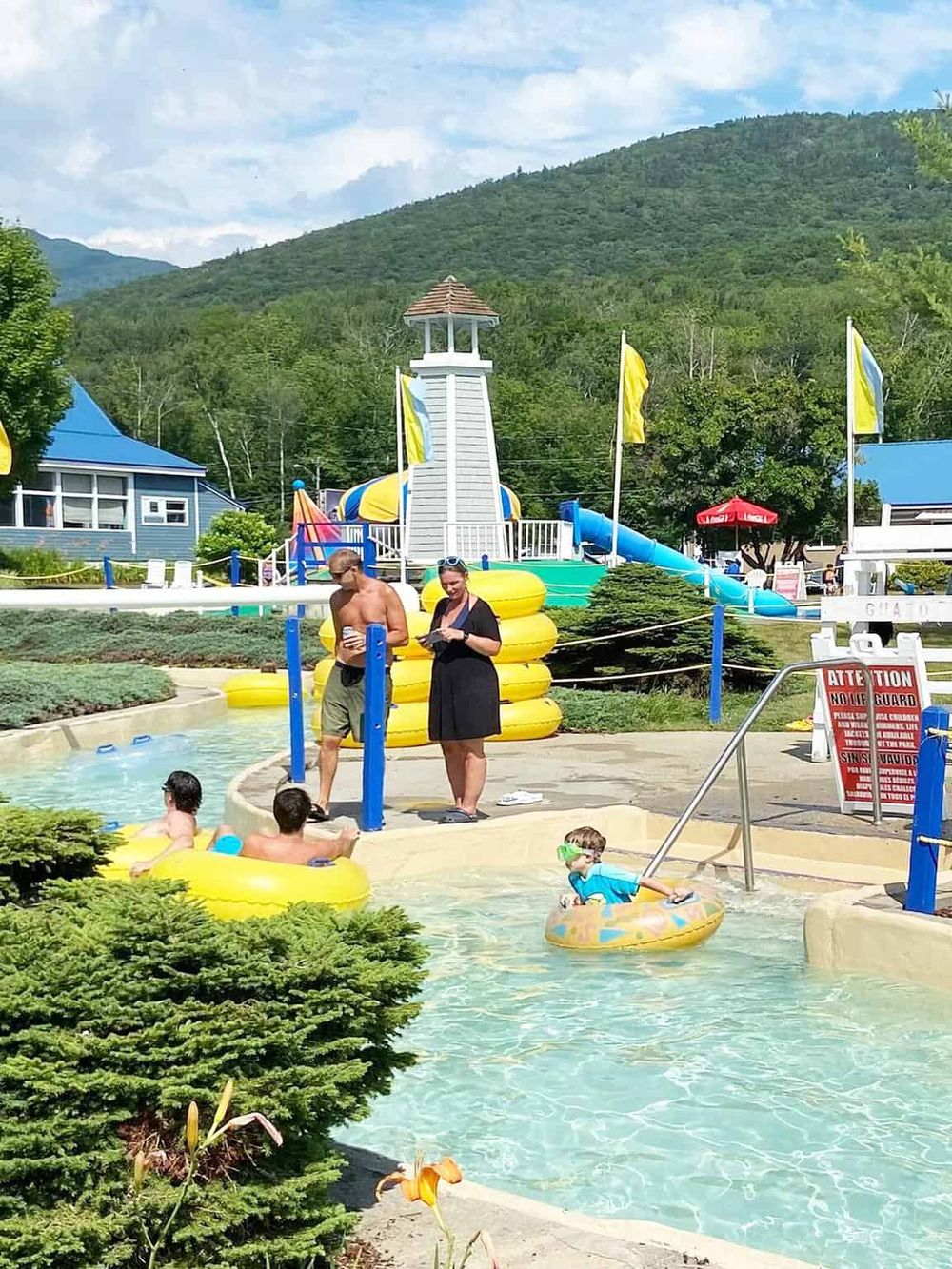 Water park with slides and children enjoying summer fun, surrounded by lush green mountains.