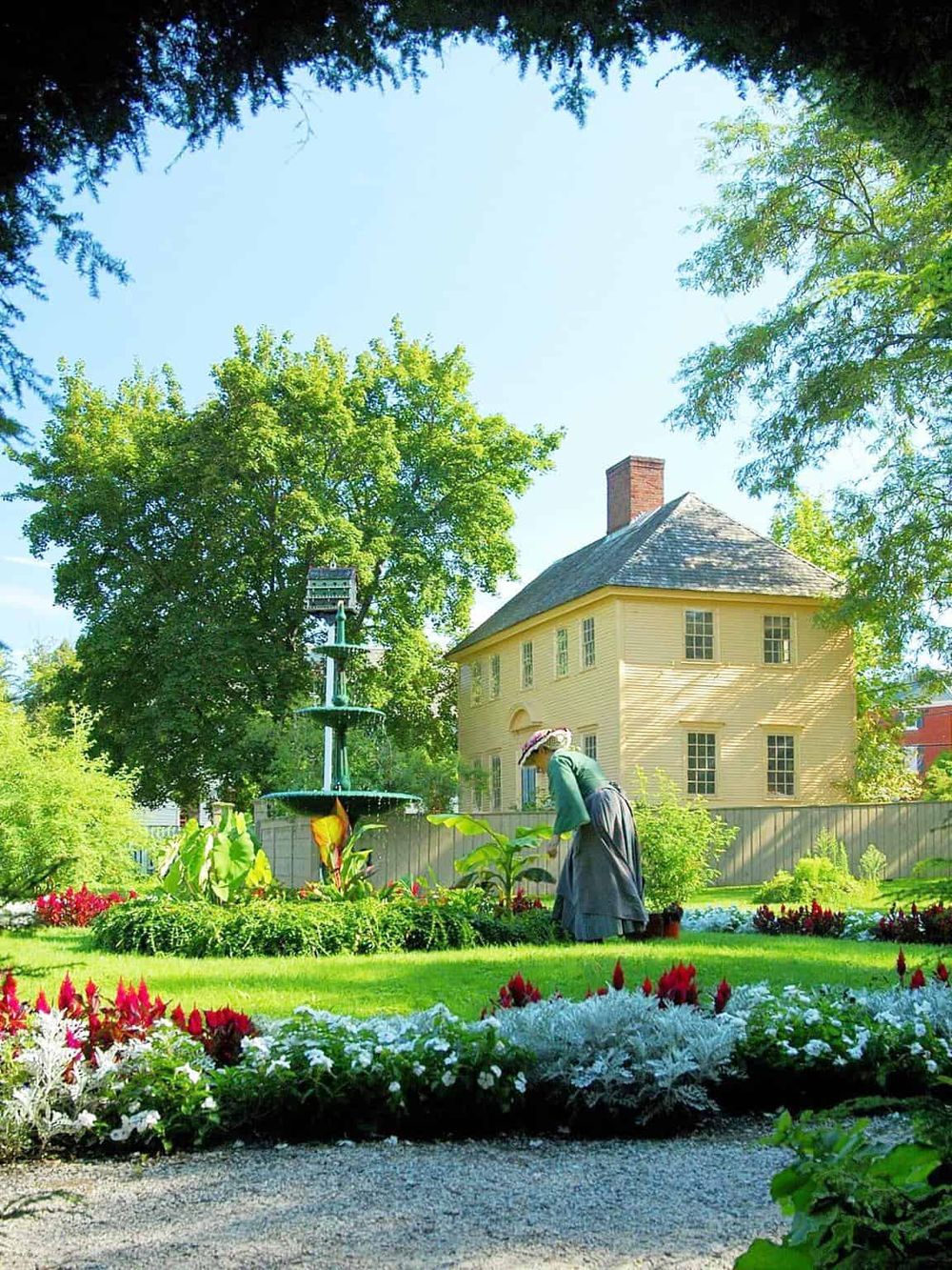 Colorful garden with a historic house and a gardener tending plants, showcasing serene outdoor spaces.