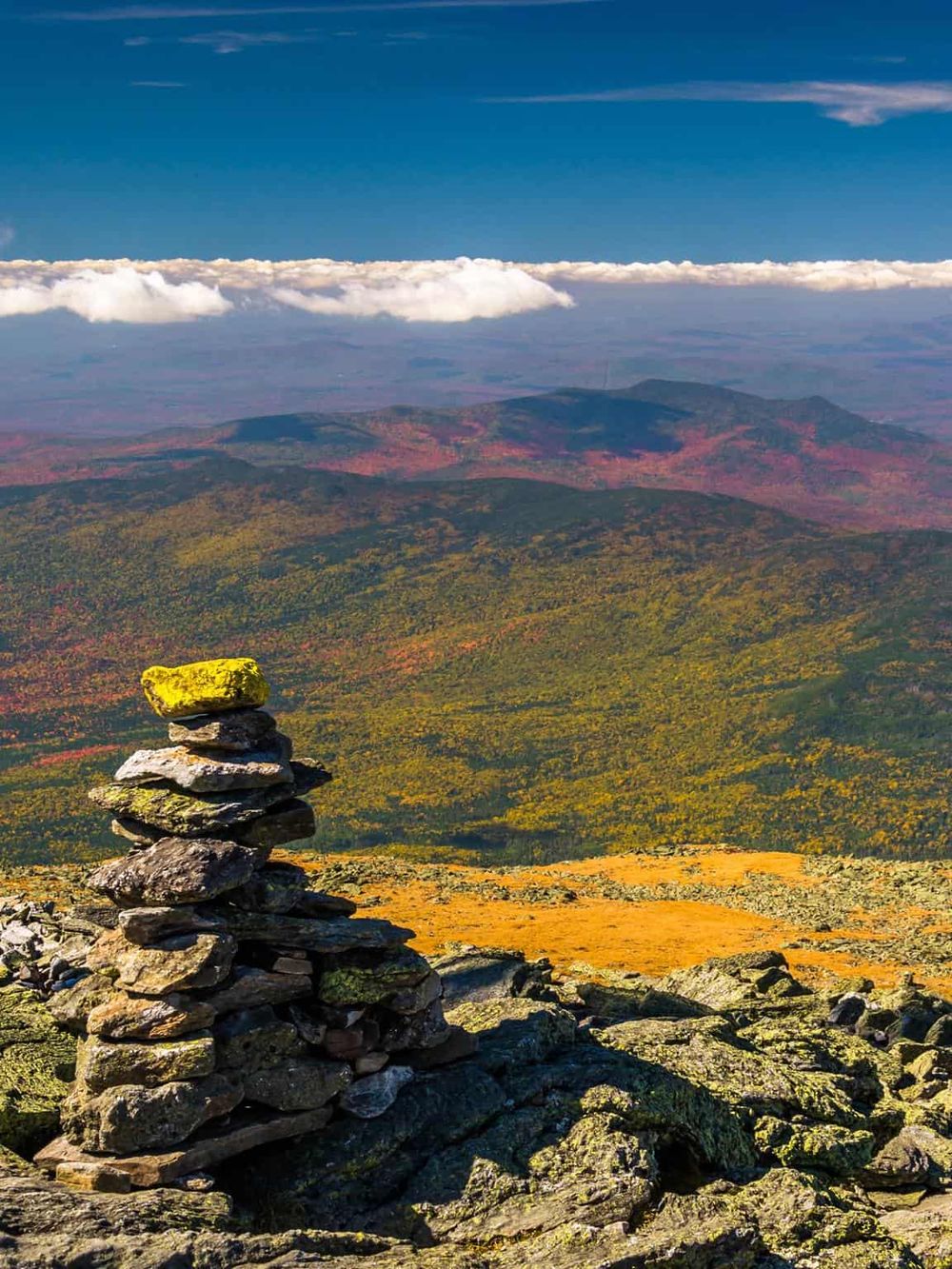 Vivid mountain landscape with cairn in foreground and scenic autumn foliage below, under a sky with clouds.