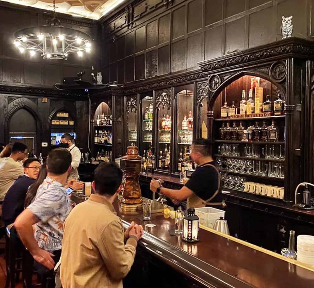 Charming bartenders serve drinks in a vintage-style bar with dark wood paneling and elegant decor.