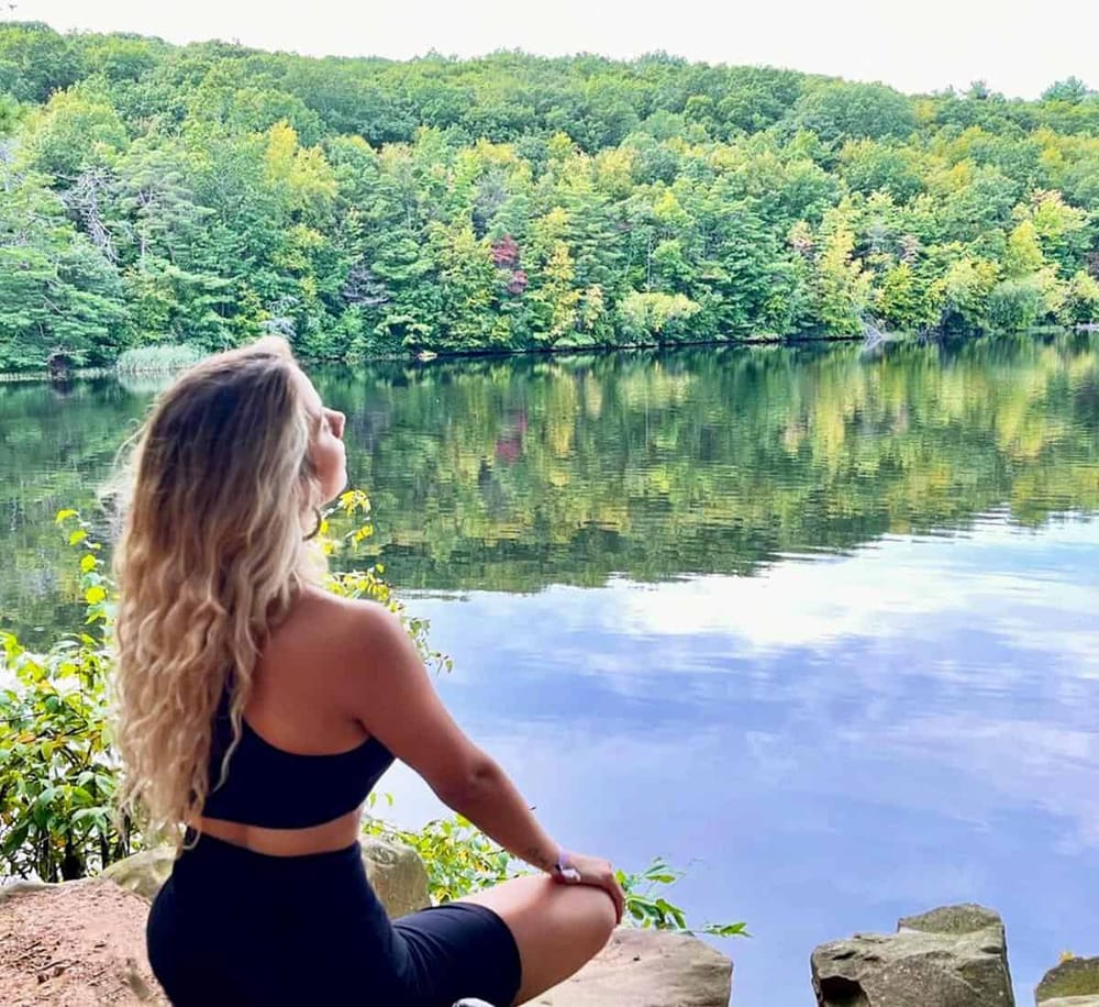Peaceful woman meditating by scenic lake surrounded by lush green trees.