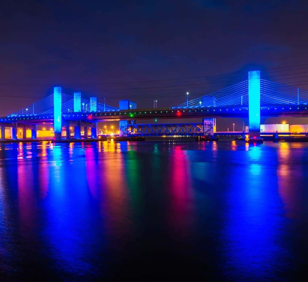 Vibrant illuminated bridge at night reflecting on water, showcasing modern architecture and cityscape views.