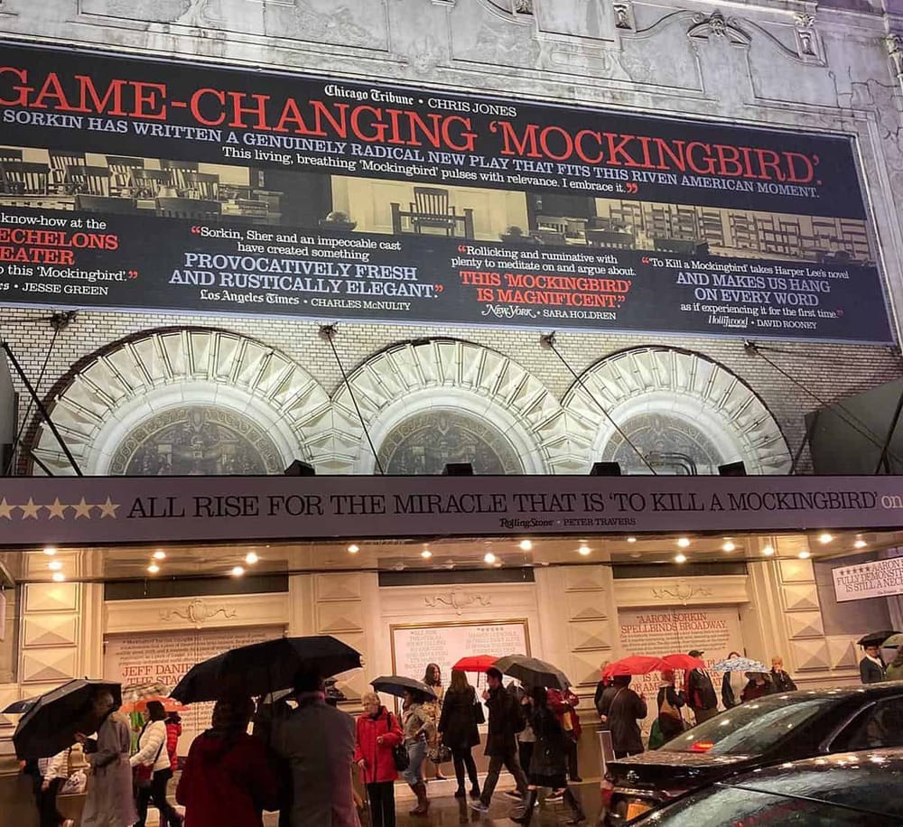 A bustling theater entrance with a large digital billboard promoting "To Kill a Mockingbird" in Times Square. People with umbrellas wait in line.