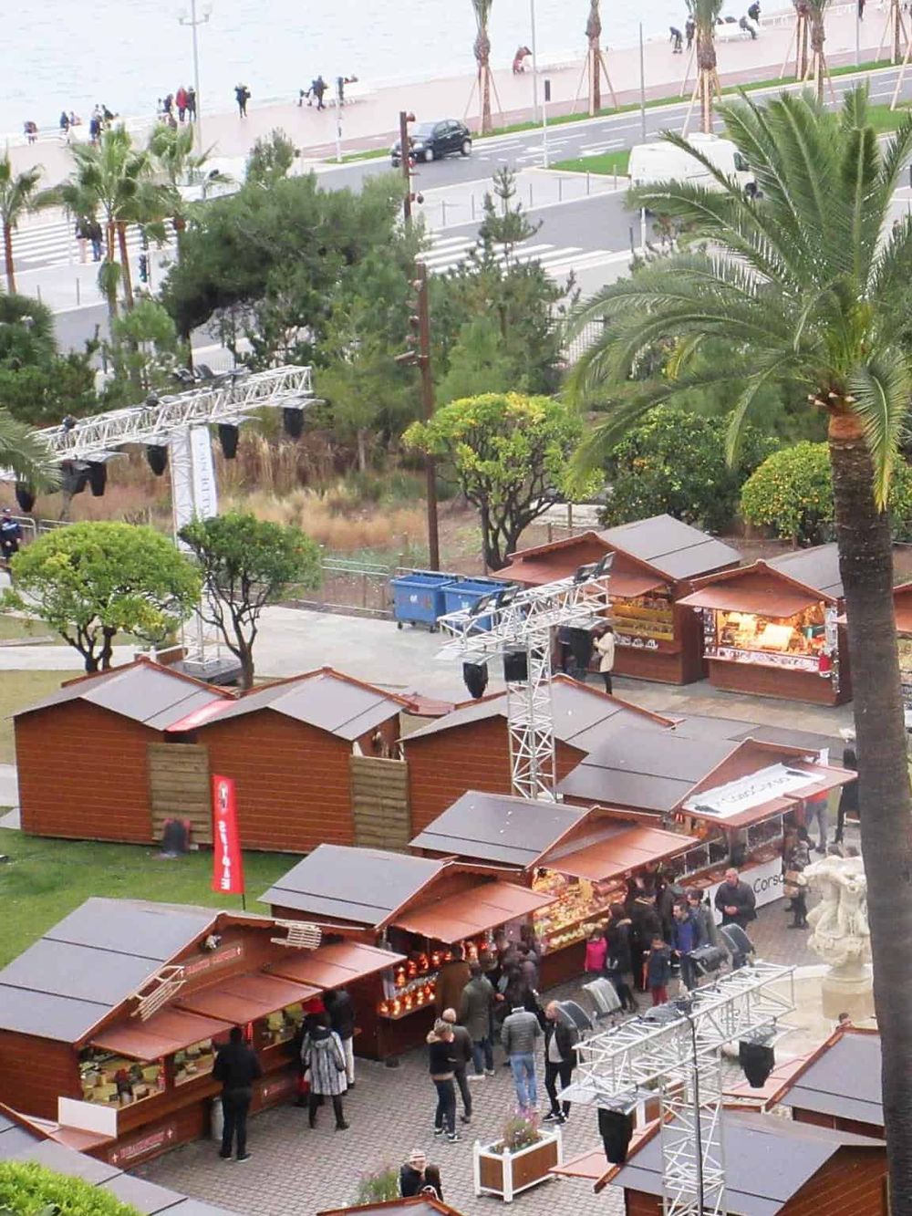 Vibrant outdoor marketplace with wooden stalls, palm trees, and visitors enjoying a scenic waterfront setting.