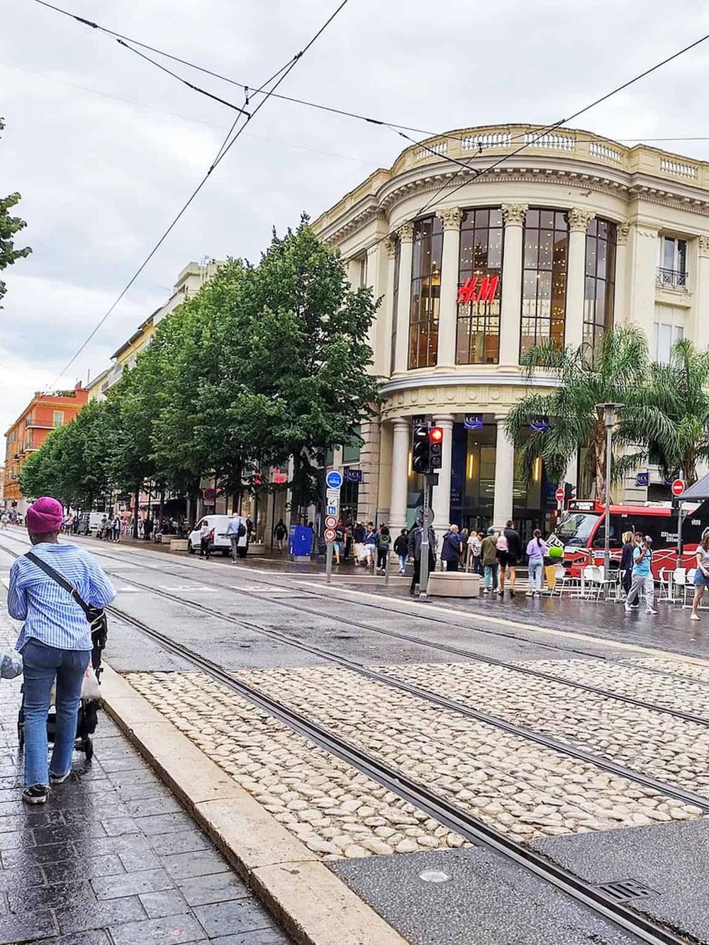 Clear view of a busy city street with tram tracks, a stylish cream-colored building with H&M store, and pedestrians at a crosswalk.