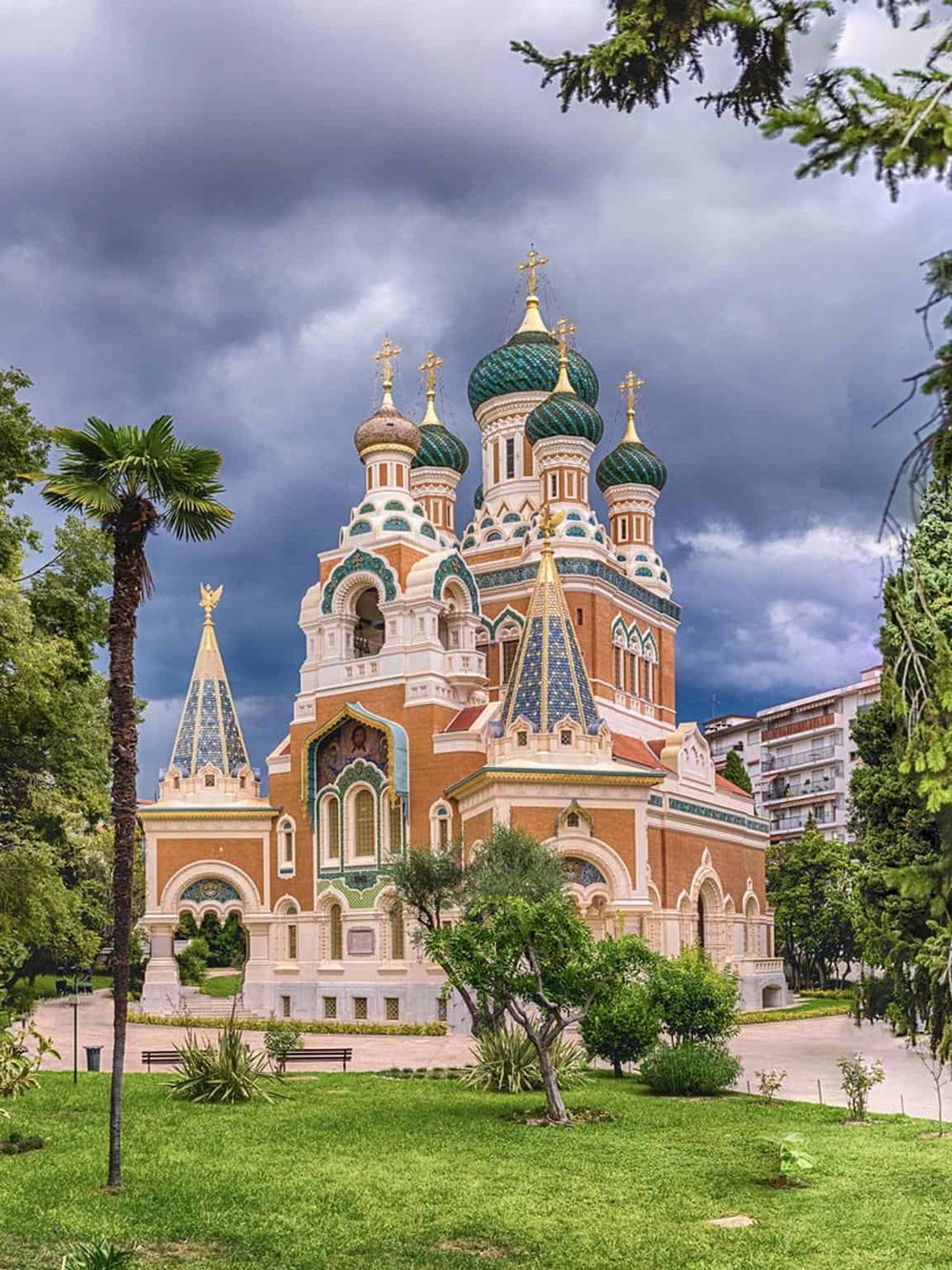 Colorful Russian Orthodox church with onion domes in a lush green park under stormy sky.