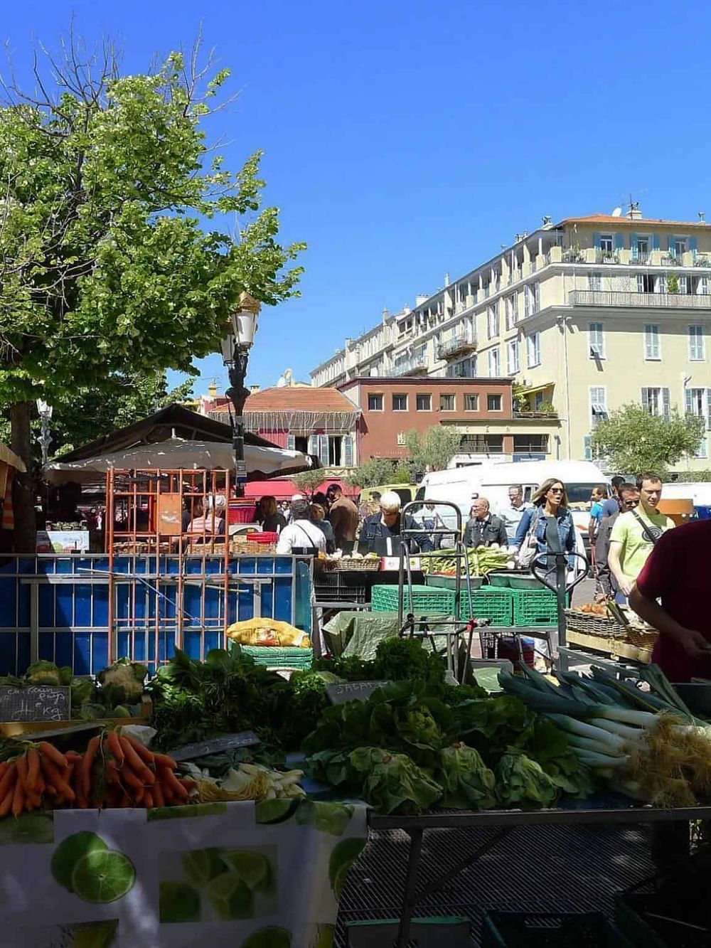 Vibrant outdoor market with fresh vegetables and bustling shoppers under a clear blue sky.