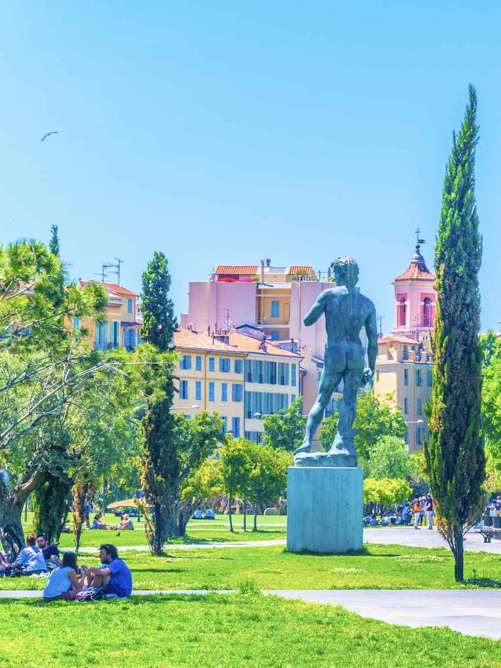 Vibrant city park featuring a classical statue, lush green trees, and colorful historic buildings in the background.