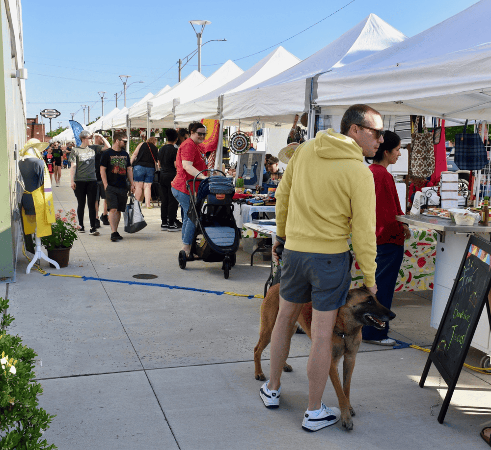 Vendors and shoppers at an outdoor market stalls with white tents on a sunny day.