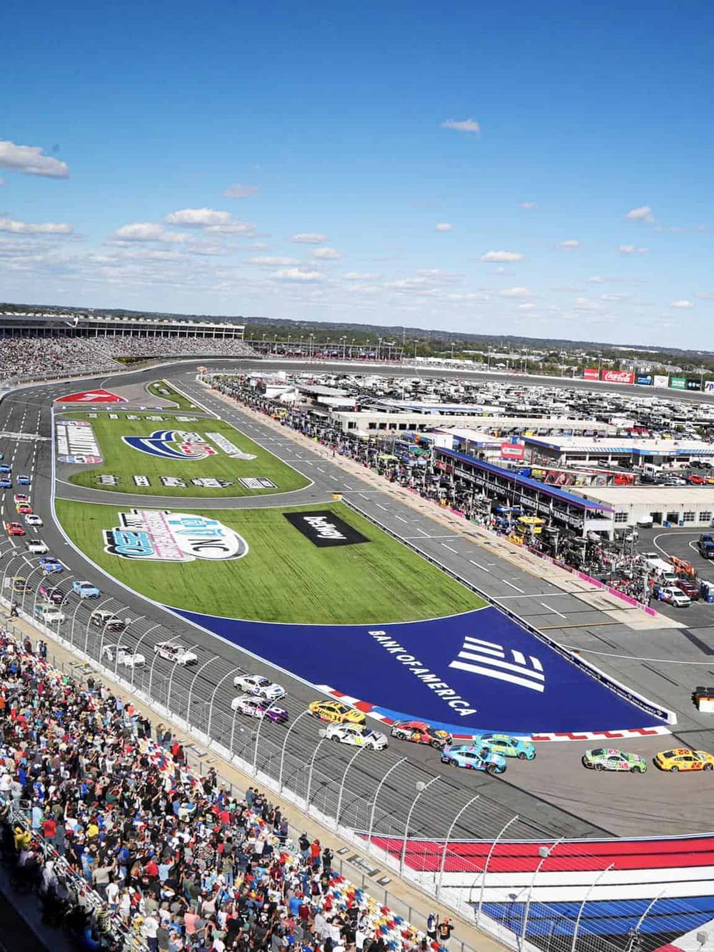 Vast NASCAR race track with cars racing, grandstands full of spectators, bright blue sky overhead.