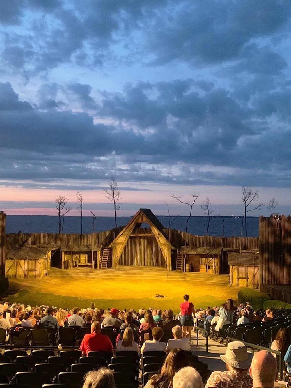 Open-air theater performance at sunset with audience watching a play on a wooden stage near the ocean.