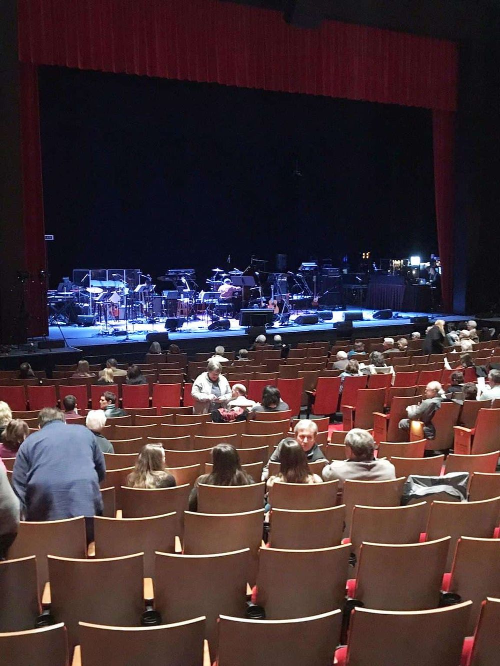 Empty theater seats before a live concert with musicians on stage and audience members arriving.