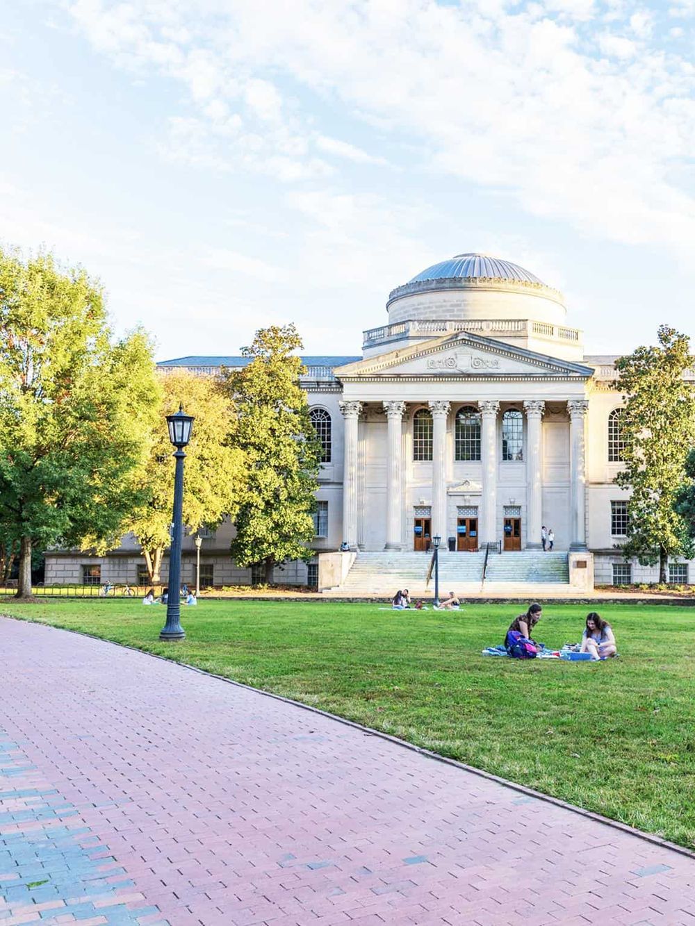 Historic university building with neoclassical architecture and surrounding greenery, students relaxing outside.