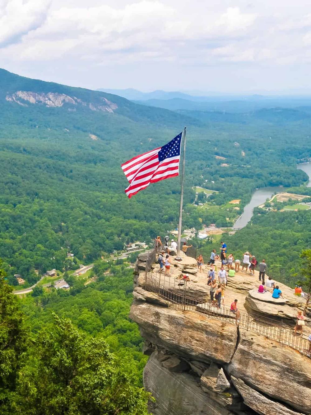 Breathtaking cliff viewpoint with American flag, overlooking lush green mountains and a river valley.