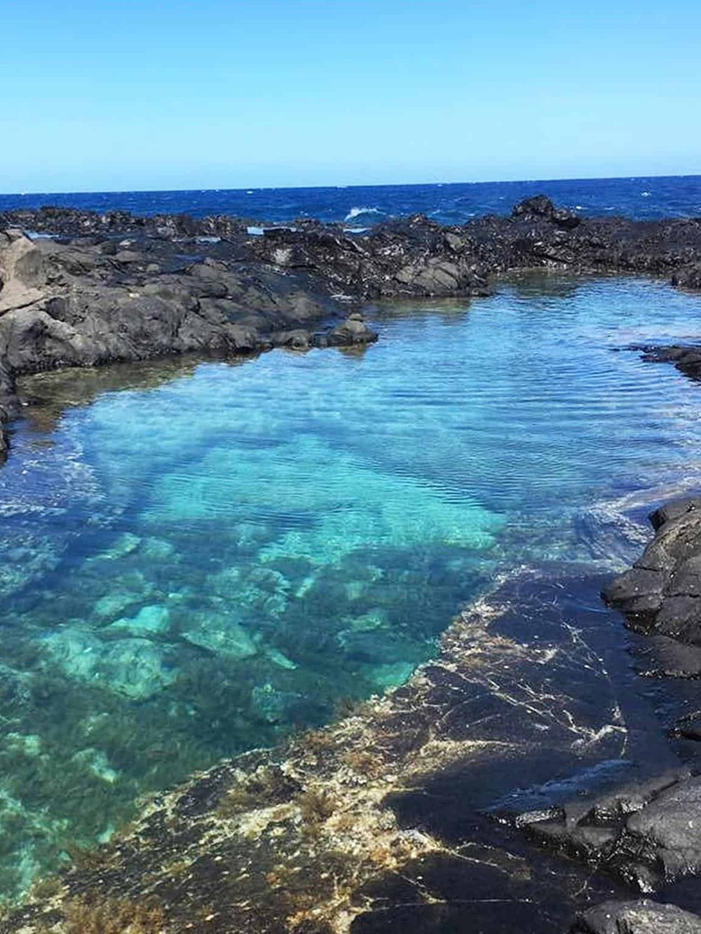 Tranquil natural swimming pool with clear blue water surrounded by black volcanic rocks on the ocean coast.