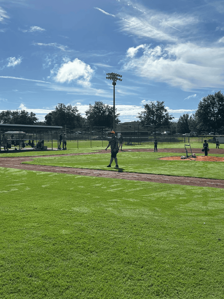 Open baseball field during daytime with players practicing and blue sky with clouds.