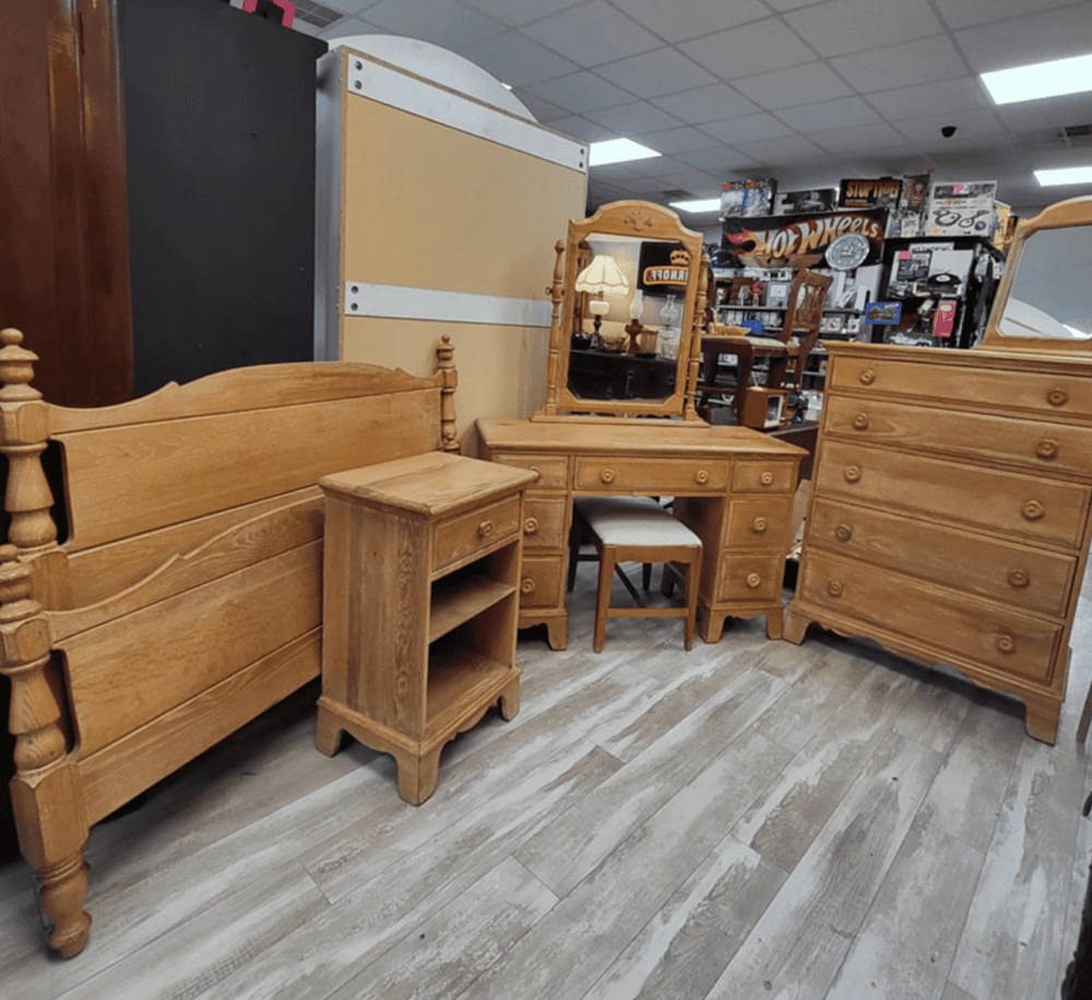 Solid wood bedroom furniture including bed, dresser, vanity, and side table at thrift store.