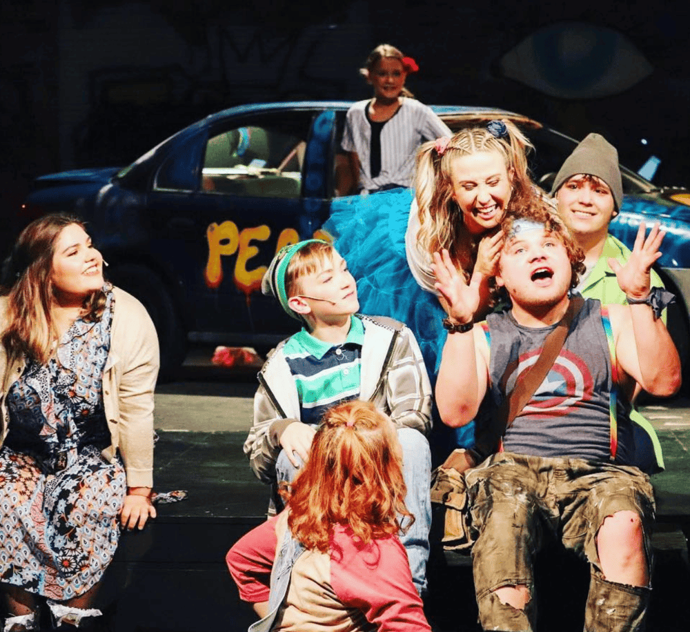 Vibrant stage performance featuring kids and an adult actor, with a colorful car prop in the background, showcasing family-friendly entertainment.