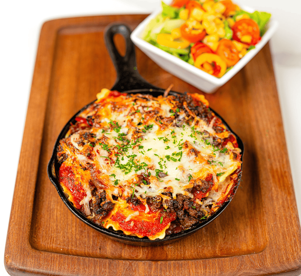 Savory skillet baked pasta with melted cheese and ground beef, served on a wooden tray with a fresh salad.