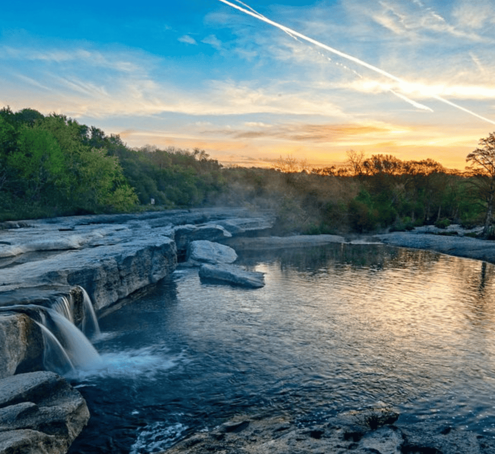 Tranquil river at sunset with lush greenery and waterfall, perfect for outdoor adventures and nature exploration.