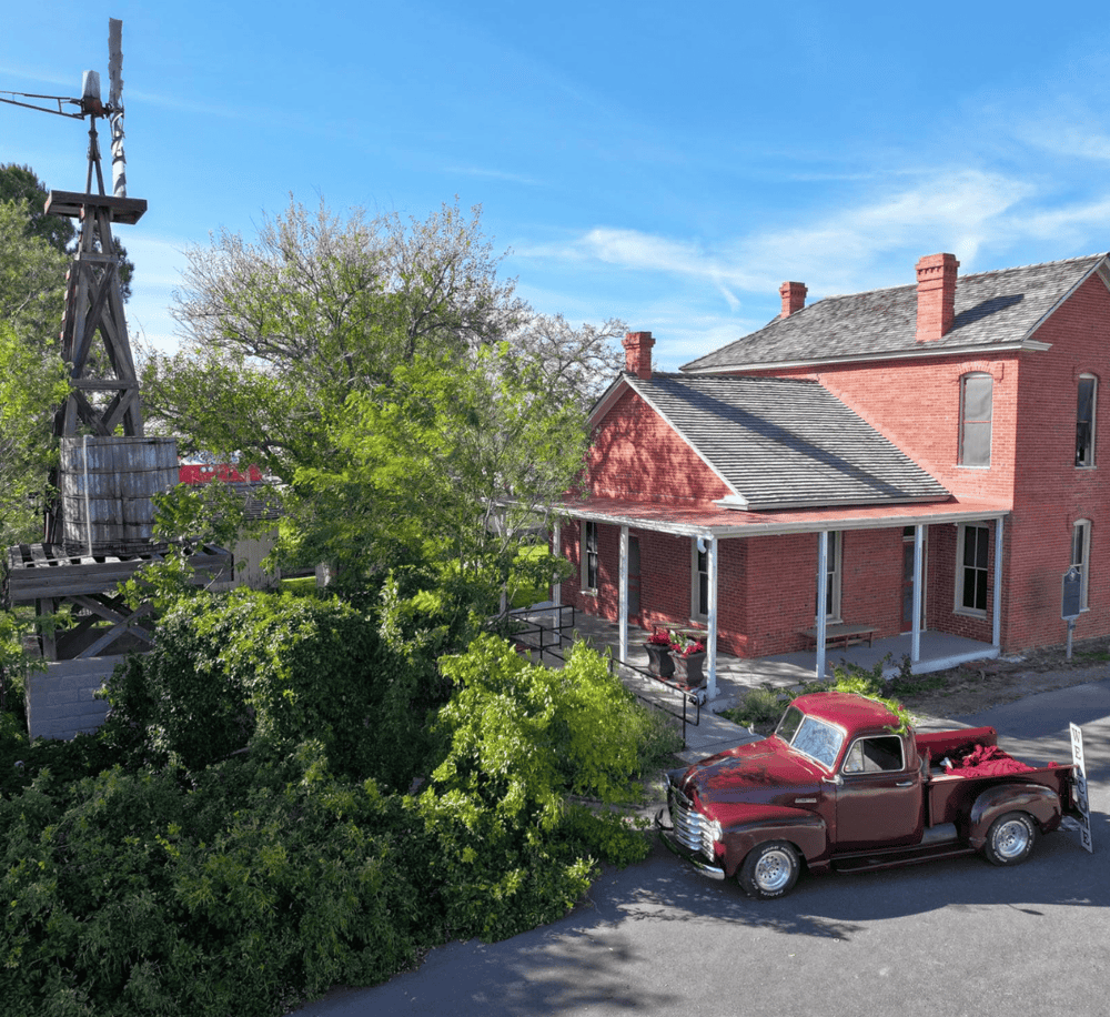 Vintage red truck parked in front of a historic brick building with a porch and lush greenery, under a clear blue sky.