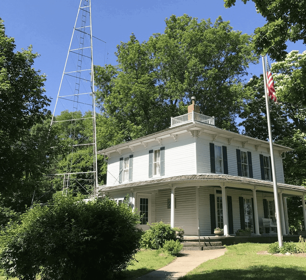 Old-fashioned house with a tall radio tower, surrounded by greenery, under a clear blue sky.