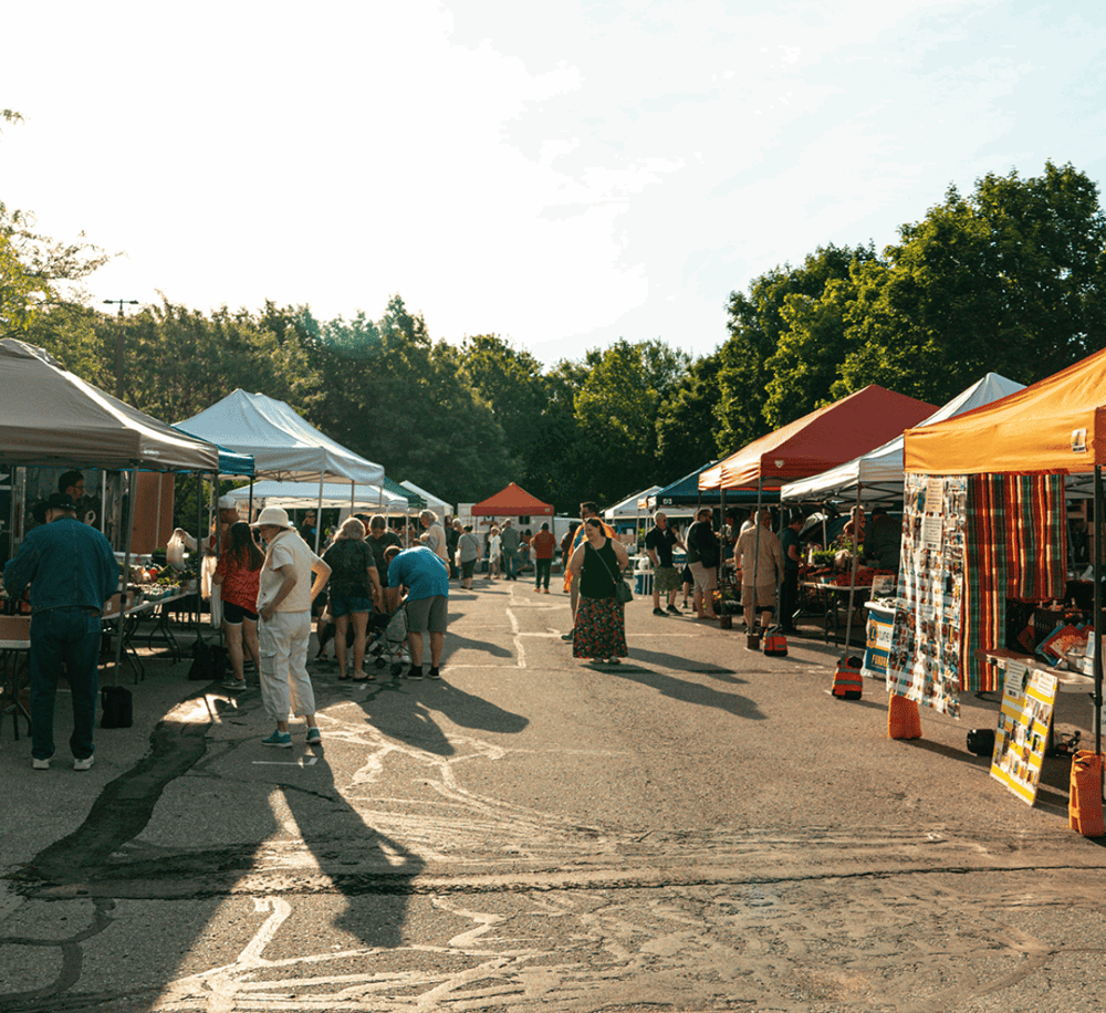 Vendors at an outdoor farmers market with colorful tents and shoppers on a sunny day.