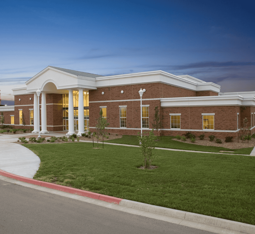 Modern community center with brick facade and white columns, surrounded by lush green landscaping.