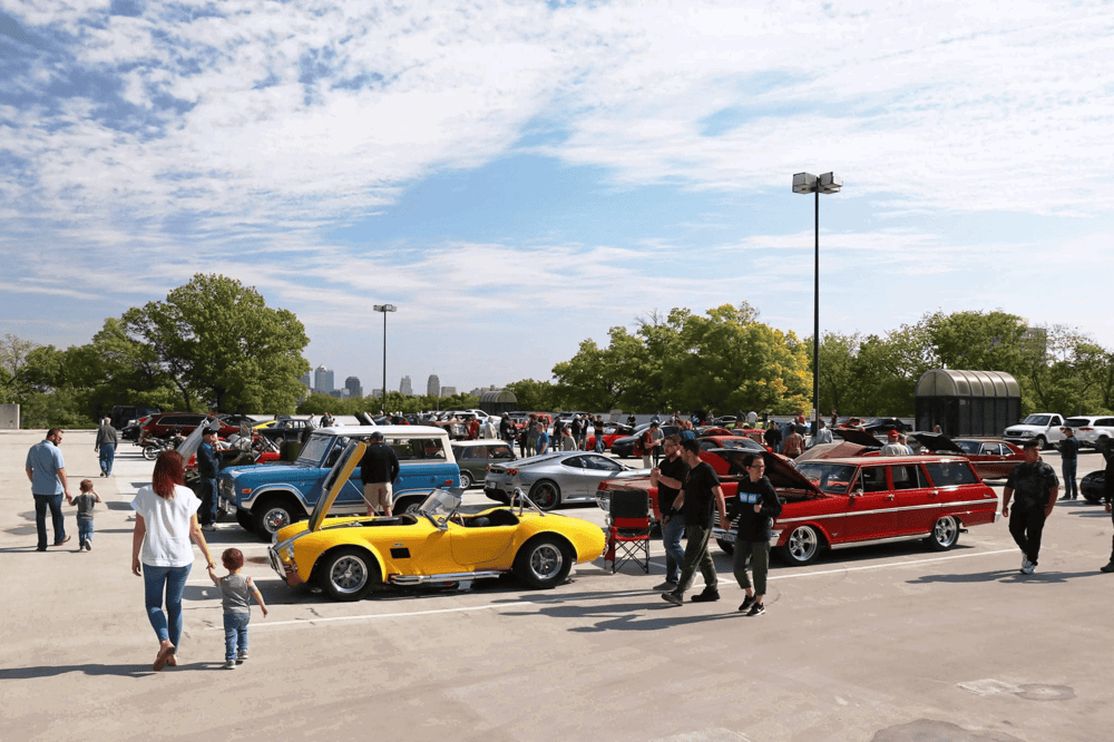 Classic car show in parking lot with vintage vehicles and family attendees.